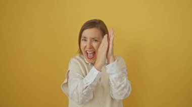 Neşeli olgun bir kadın beyaz bluz giyerek sarı izole arka plan üzerinde el çırpıyor.