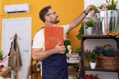 Middle age man florist touching plant holding clipboard at flower shop