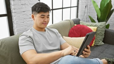 Genç Asyalı bir adam, modern bir dairede rahat bir koltukta tablet okurken rahatlıyor..