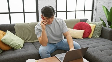 Genç Asyalı bir adam modern bir oturma odasında dizüstü bilgisayarıyla çalışırken telefonunu kullanıyor..