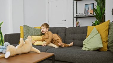 Gri bir kanepede uzanan gülümseyen çocuk rahat bir oturma odasında renkli yastıklarla