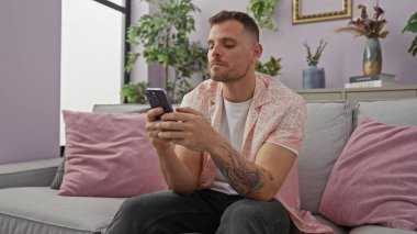Modern oturma odasında akıllı telefon kullanan dövmeli yakışıklı bir adam.
