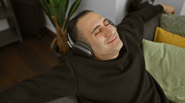 Rahat İspanyol adam, rahat bir oturma odasında kulaklıkla müzik dinliyor.