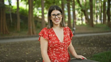 Şehir parkında poz veren neşeli, çekici İspanyol kadın. Güzel, yetişkin esmer, utanmadan gülümseyen, doğal neşe ve güven saçan, gözlüklü.
