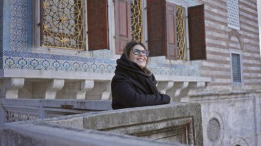 Gözlüklü, sıcacık giyinmiş, gülümseyen bir kadın, Osmanlı mimarisinin yer aldığı tarihi İstanbul Sarayı 'nı ziyaret ediyor..