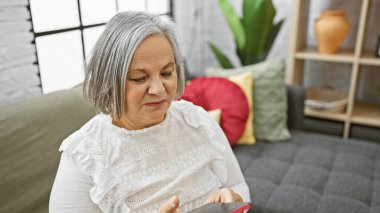 Rahat bir oturma odasında akıllı telefonuyla meşgul olan olgun bir kadın, bağlantıyı ve boş zamanlarını yansıtıyor..