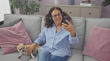 Orta yaşlı, gülümseyen, kulaklıklı bir İspanyol kadın evcil köpeğiyle evde bir kanepede selfie çekiyor..
