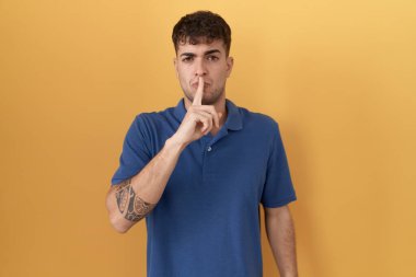 Sarı arka planda dikilen genç İspanyol adam parmakları dudaklarında sessiz olmak istiyor. sessizlik ve gizli konsept. 