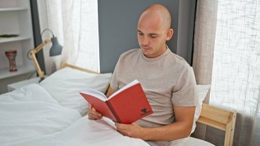Young hispanic man reading book sitting on bed at bedroom