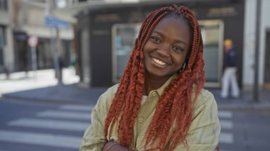 Şehir caddesinde duran kırmızı örgülü neşeli, Afro-Amerikan bir kadın.