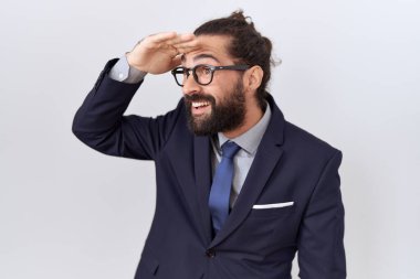 Hispanic man with beard wearing suit and tie very happy and smiling looking far away with hand over head. searching concept. 
