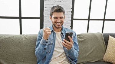 Sakallı, genç, Latin kökenli yetişkin adam, oturma odasında akıllı telefonuyla başarısını kutluyor..