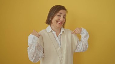 Gülümseyen orta yaşlı bir kadın sarı arka planda kendini işaret ediyor.