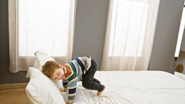 İyi aydınlatılmış bir yatak odasında beyaz bir yatakta yuvarlanan çizgili gömlekli beyaz çocuk.