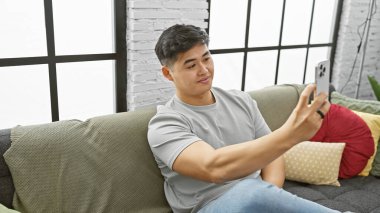 Yakışıklı Asyalı adam modern oturma odasında akıllı telefonuyla selfie çekiyor.