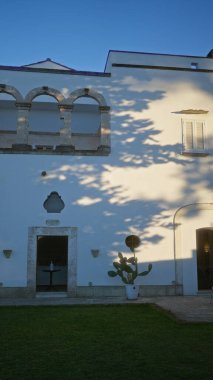 Güneş ışığıyla aydınlatılan taştan kemerli ve yeşil çimenli zarif beyaz bina, puglia, İtalya 'da yer alıyor, geleneksel Avrupa mimarisi ve açık hava sükuneti sergiliyor..