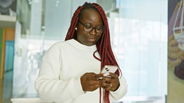 Parlak, modern bir ofiste akıllı telefon kullanan örgülü Afrikalı Amerikalı bir kadın.