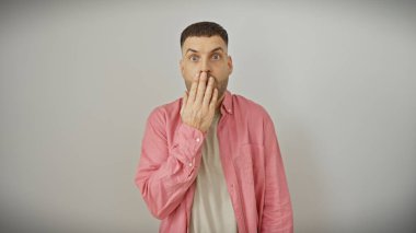 Pembe tişörtlü, sakallı, beyaz arka planda ağzını kapatan şaşırmış genç bir adam.