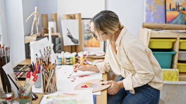 Sanat stüdyosundaki masanın üzerine resim çizen genç sarışın kadın sanatçı.
