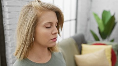 Düşünceli, kısa saçlı genç bir kadın modern bir oturma odasında dinleniyor, rahat bir kanepesi ve çiçekleri var.