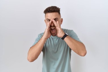 Hispanic man with beard standing over white background shouting angry out loud with hands over mouth 