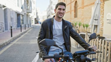 Handsome hispanic man in leather jacket smiling on motorcycle in sunny urban street scene. clipart