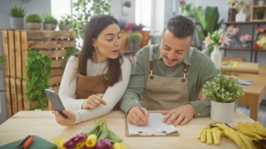 Telefonlu bir kadın ve not alan bir adam, ikisi de çiçekçinin ortasında önlük takıyor..