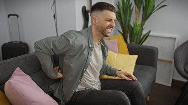 Genç İspanyol bir adam, modern bir oturma odasındaki bir kanepede oturup eski sayıları ima ederken acı içinde kıvranıyor..