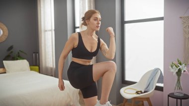 Aktif giyinen genç bir kadın modern bir yatak odasında doğal ışıkla esniyor..