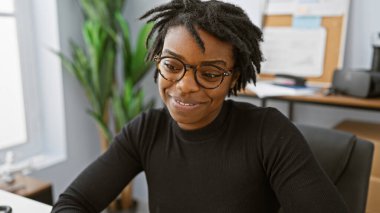 Rasta takan, gözlüklü, ofisteki bir iş yerinde oturan gülümseyen siyah bir kadın..