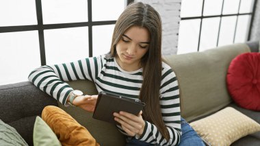 İçerideki koltukta tablet kullanan Latin kökenli genç bir kadın. Modern, rahat bir iç mekanı var..