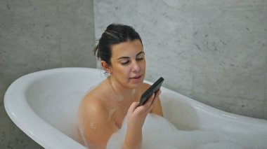 Genç bir kadın, çağdaş bir banyo ortamında akıllı telefonunu karıştırırken evinde rahatlatıcı bir köpük banyosunun keyfini çıkarıyor..