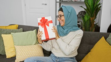 Sırıtan tesettürlü kadın, elinde hediyeyle kanepede oturuyor.