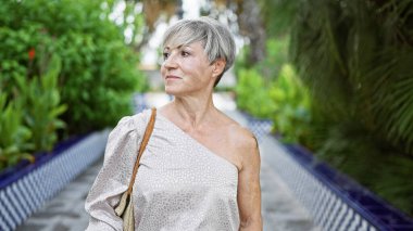 Kısa gri saçlı, olgun İspanyol bir kadın açık havada yeşil yemyeşil bir bahçede düşünceli bir şekilde yürüyor..