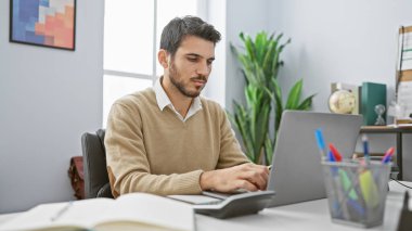 Modern ofis ortamında dizüstü bilgisayarda çalışan yakışıklı İspanyol bir adam. Odaklanma ve profesyonellik gösteriyor..
