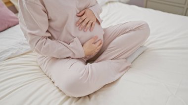 Genç bir İspanyol kadın yatakta oturur, hamile karnını rahat bir yatak odasında sarmalar, hamilelik sükunetini hatırlatır..