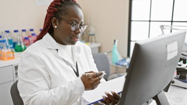 Laboratuvarda çalışan, laboratuvar önlüğü ve gözlüklü Afro-Amerikan kadın..
