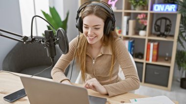 Radyo stüdyosunda kulaklıkla podcast yapan gülümseyen genç bir kadın.