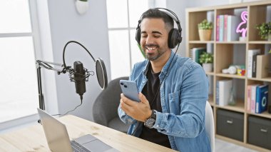 Telsiz stüdyosunda çalışan, elinde akıllı telefon tutan ve kulaklık takan, sakallı, gülümseyen bir İspanyol..