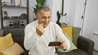 Middle-aged hispanic man playing video game in living room, joyful expression, home interior. clipart