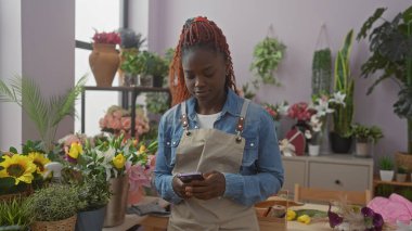 Genç bir Afrikalı Amerikalı kadın çeşitli bitkilerle dolu renkli bir çiçekçide akıllı telefon kullanıyor.