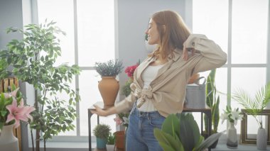 Neşeli kızıl saçlı bir kadın yeşil bitkiler ve doğal ışıklarla çevrili parlak bir odada giyinir..