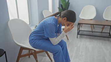 A distressed young woman in blue scrubs sits in a bright hospital room, covering her face with her hands. clipart