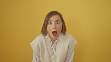 Şaşırmış orta yaşlı bir kadın sarı arka planda, kısa saçlı, geniş gözlü ve ağzı açık bir kadın..