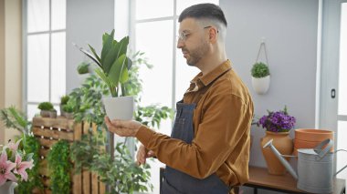 Önlüklü İspanyol adam, arka planda tencere ve saksı bulunan ayışığı çiçekçisindeki bitkileri değerlendiriyor..