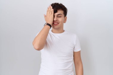 Gündelik beyaz tişört giyen, bir gözünü eliyle kapatan, yüzünde kendine güvenen bir gülümseme ve sürpriz duygular taşıyan genç bir adam.. 