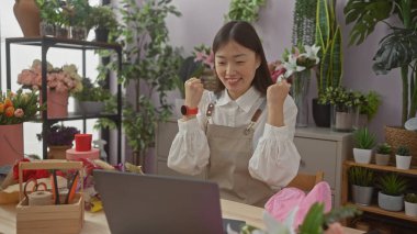 Çiçekçi dükkanında başarıyı kutlayan heyecanlı Asyalı kadın bitkiler ve dekorlarla çevrili.