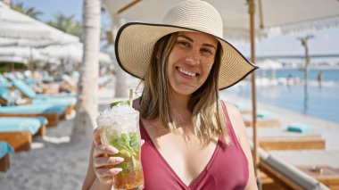Güneşli bir günde havuz başında şapkalı ve mayo giymiş gülümseyen bir kadın kokteyl içiyor..
