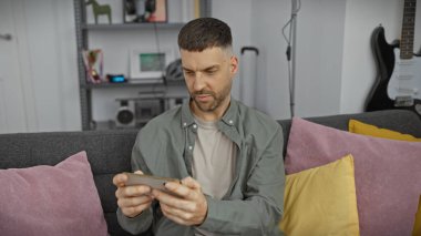 İspanyol adam rahat, modern oturma odasında gitar çalan akıllı telefona odaklanmış.