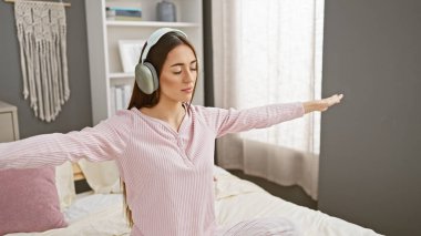 A woman in a pink sweater and headphones stretching on a bed in a cozy bedroom with neutral colors. clipart